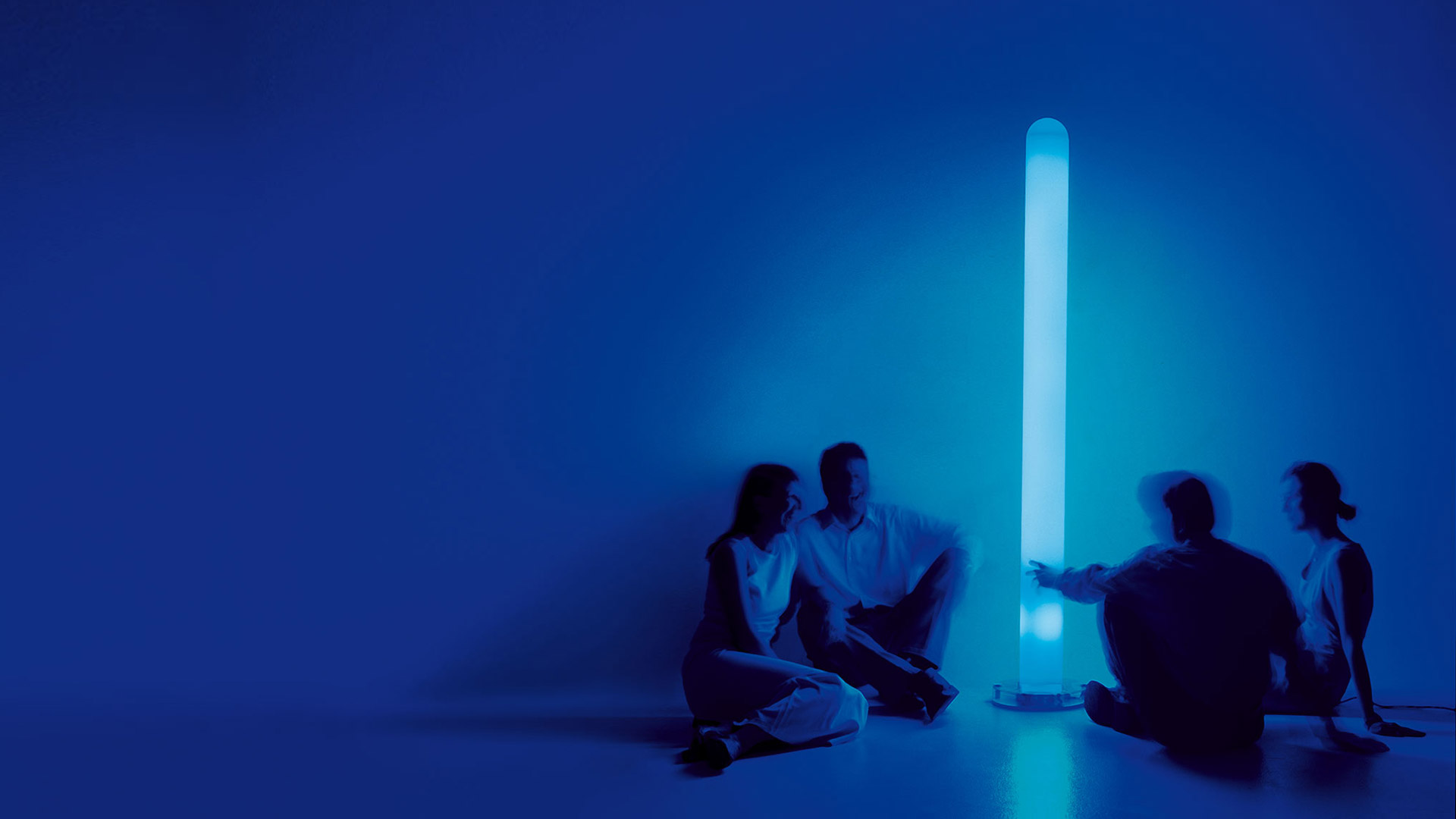 Four people sitting around Artemide Metacolor tall cylindrical floor lamp glowing in blue light in a blue-lit room