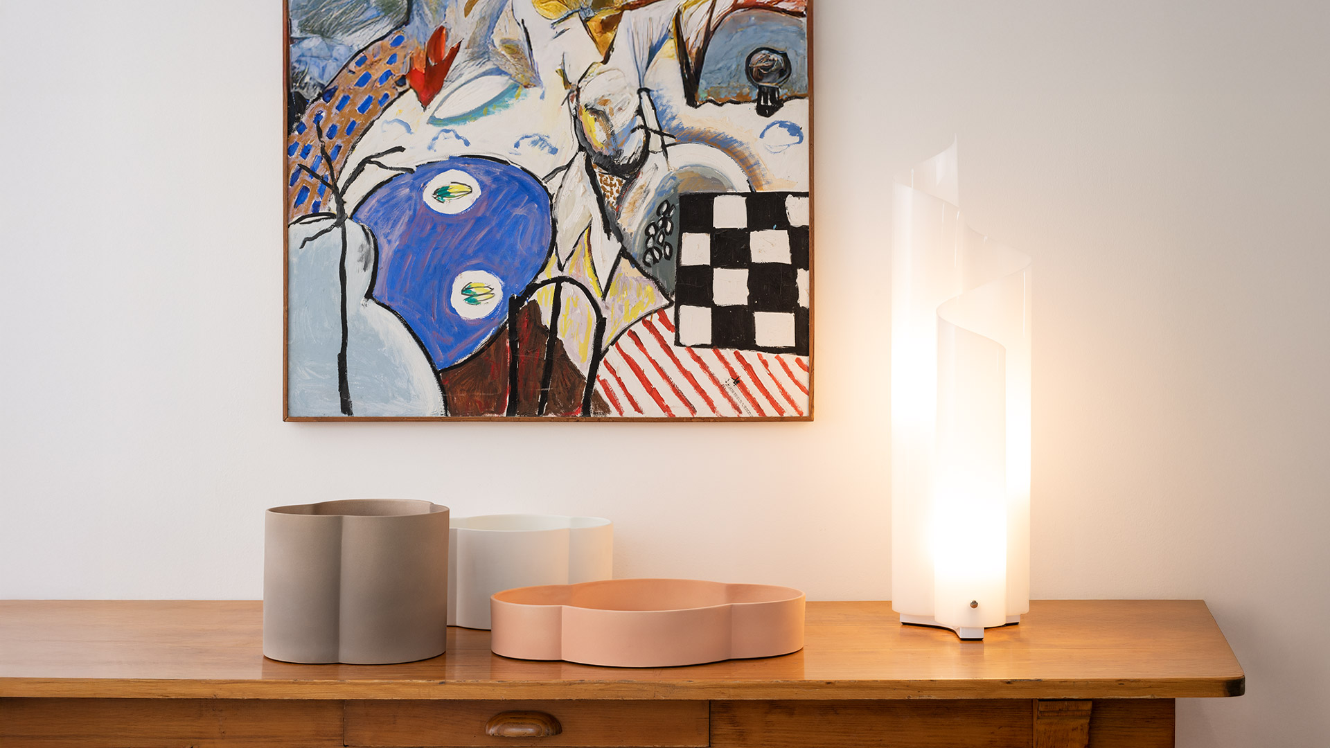 Artemide Mezzachimera table lamp with white opal diffuser glowing on a wooden sideboard, beside decorative objects and a colorful painting