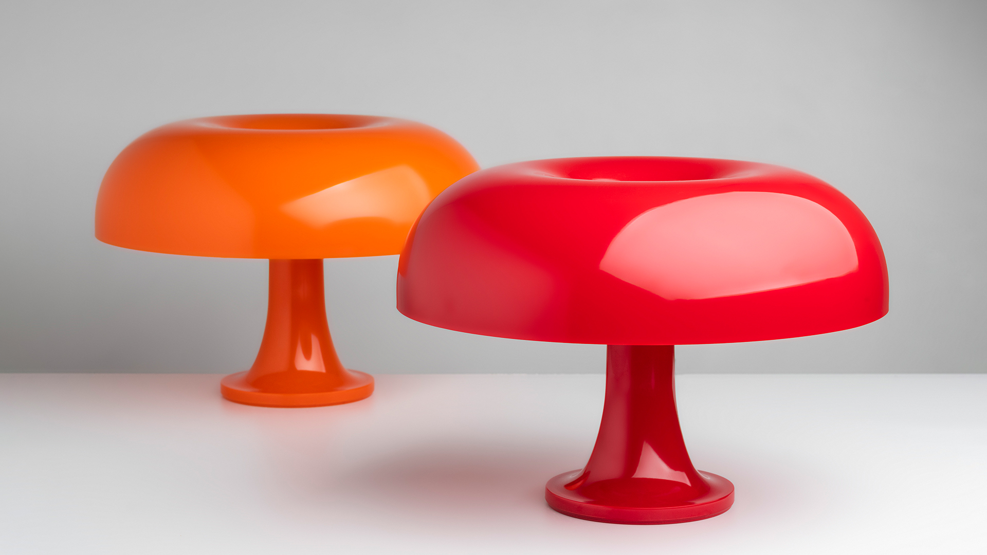 Two Artemide Nessino table lamps with mushroom-shaped shades in deep red, displayed on tiered cylindrical pedestals against red velvet curtains