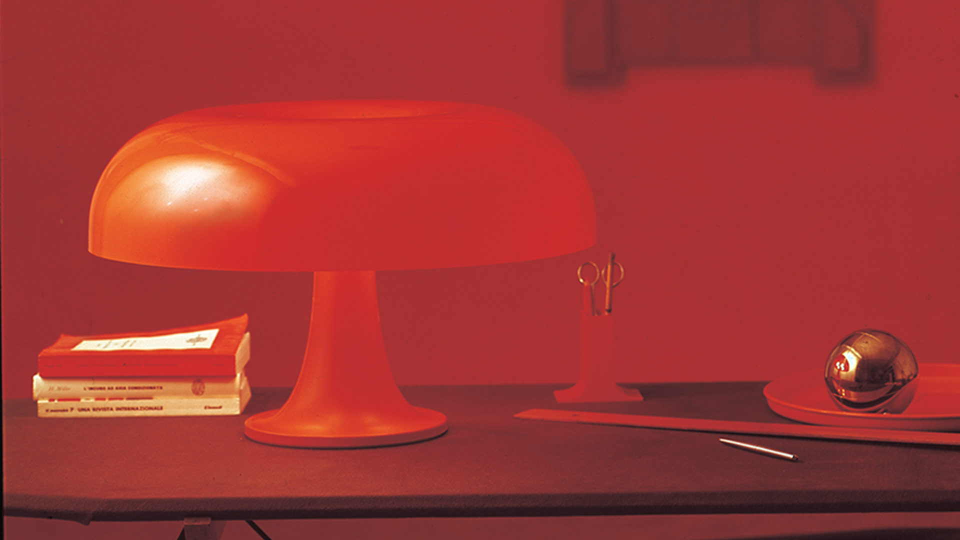 Artemide Nesso table lamp in red, featuring iconic mushroom-shaped shade on a trumpet base, glowing warmly on a white pedestal