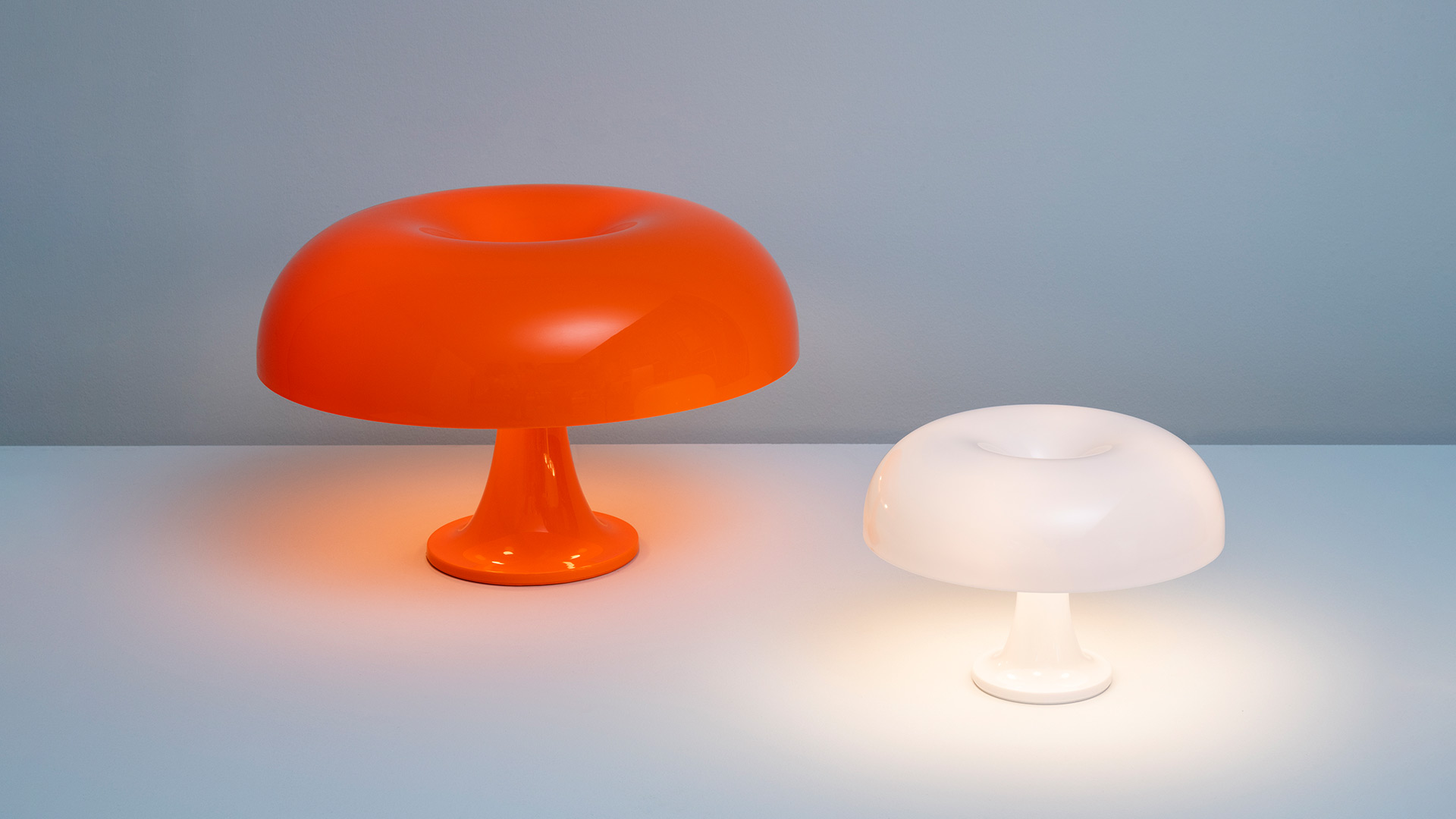 Two Artemide Nesso table lamps: large orange mushroom-shaped lamp on left, smaller white illuminated version on right