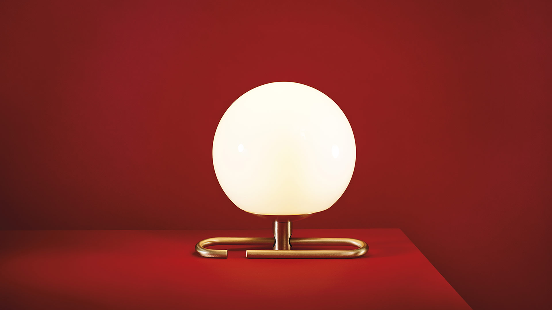 Artemide NH 1217 table lamp with opal white glass sphere and brass base, glowing warmly on a shelf with books and decorative sculptures