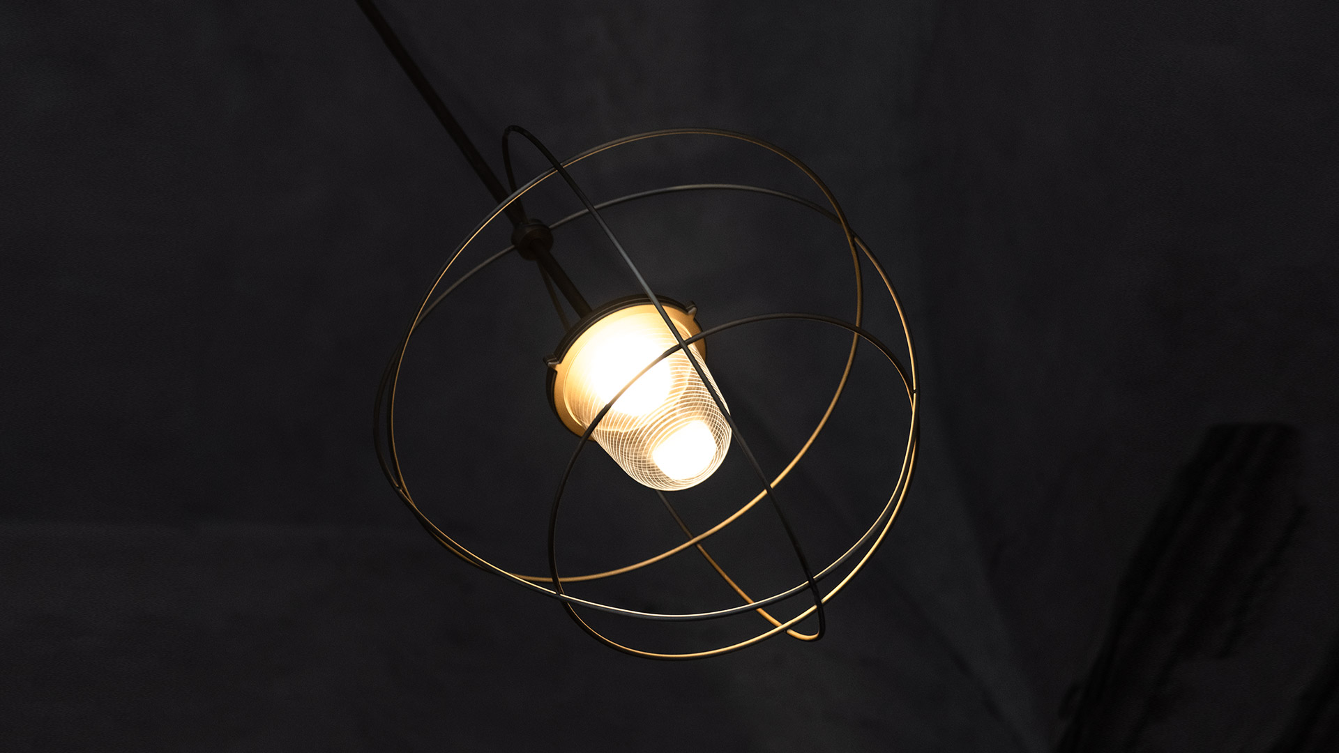 Nottola pendant lamp with gold metal wire orbital rings framing a glowing frosted bulb, suspended from black cord against dark background