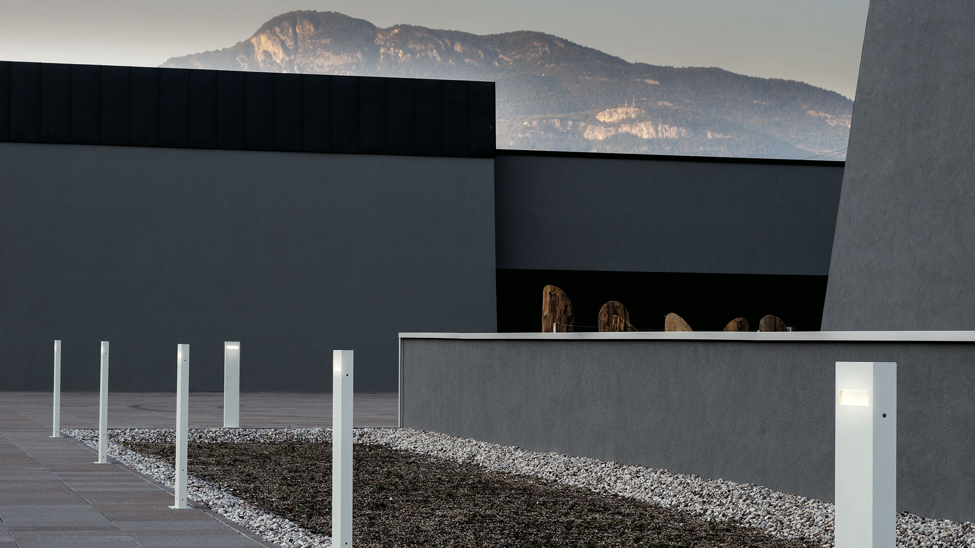 Two white rectangular Artemide Nuda outdoor bollard lights of different heights glowing softly in a grassy field at dusk, with a rooster nearby