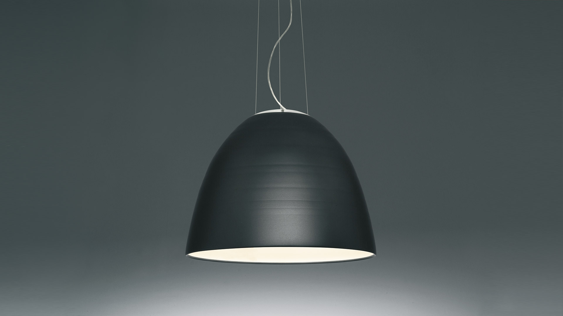 Indoor equestrian arena with multiple Artemide Nur 1618 pendant lamps on sloped white ceiling, illuminating sandy riding floor with horse and riders
