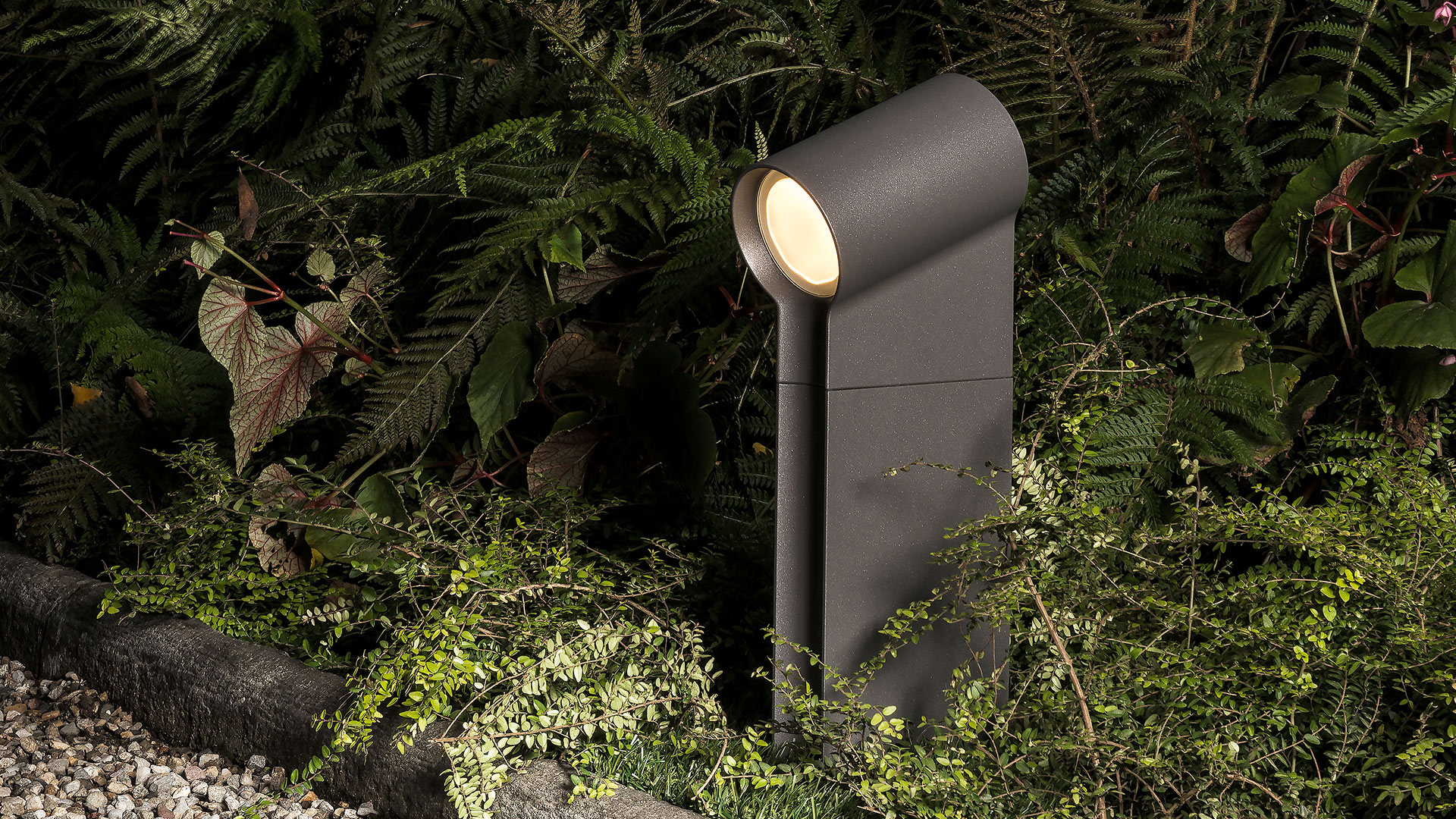 Artemide Oblique outdoor bollard light in dark grey, with angled LED head illuminating a lush garden at night