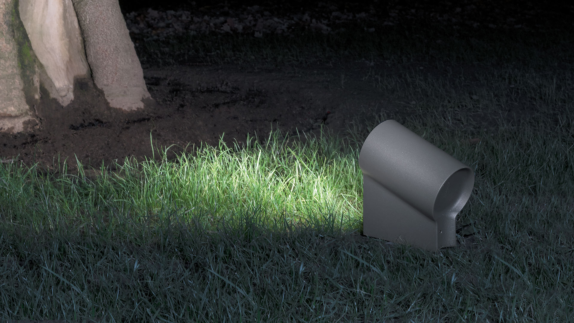 Artemide Oblique outdoor spotlight in dark grey, ground-mounted on grass, casting focused upward light on a tree base at night