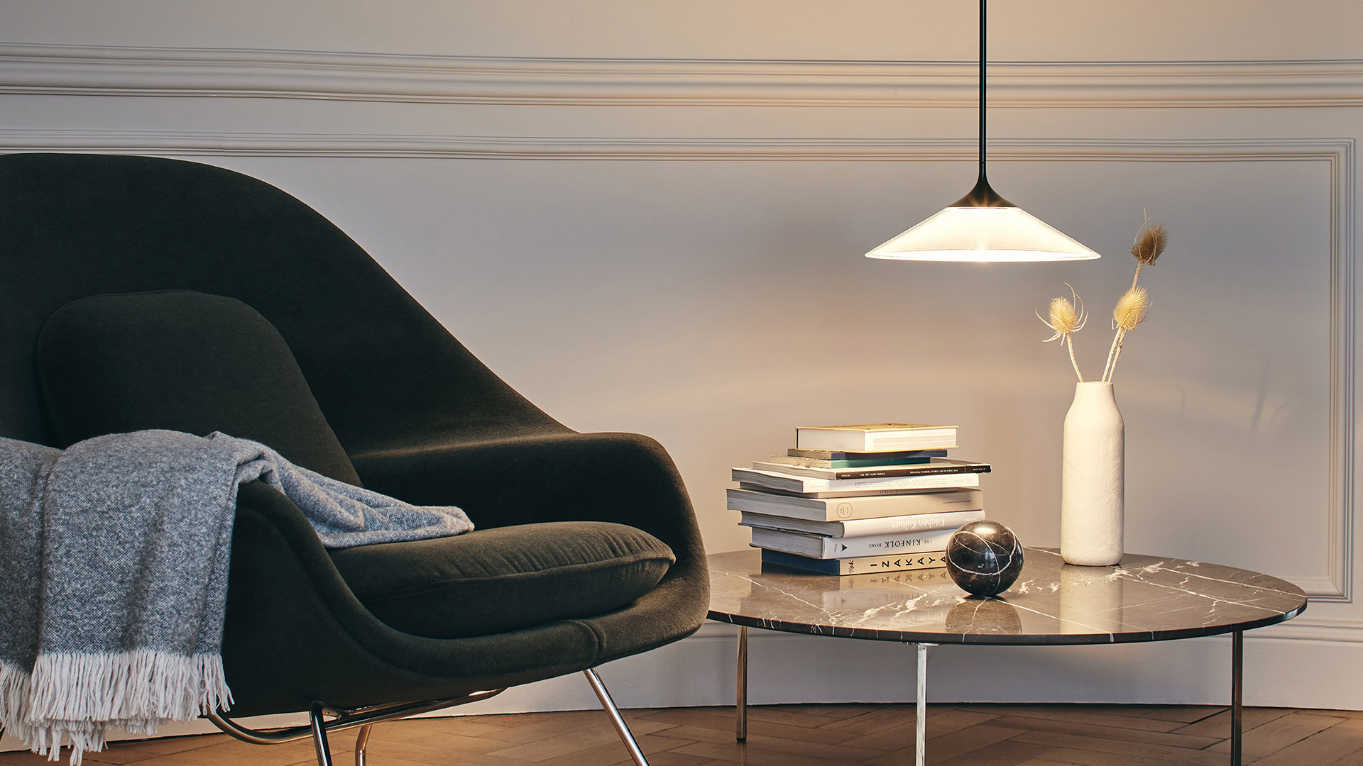 Artemide Orsa pendant lamp with white opal shade illuminates a marble side table with books and vase, beside a dark velvet armchair