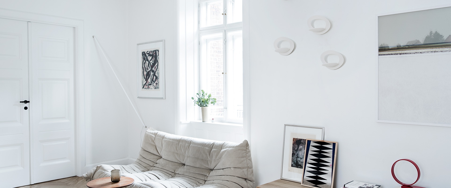 Minimalist white living room with a grey sofa, wooden table, and three Artemide Pirce Parete wall lamps mounted on the wall