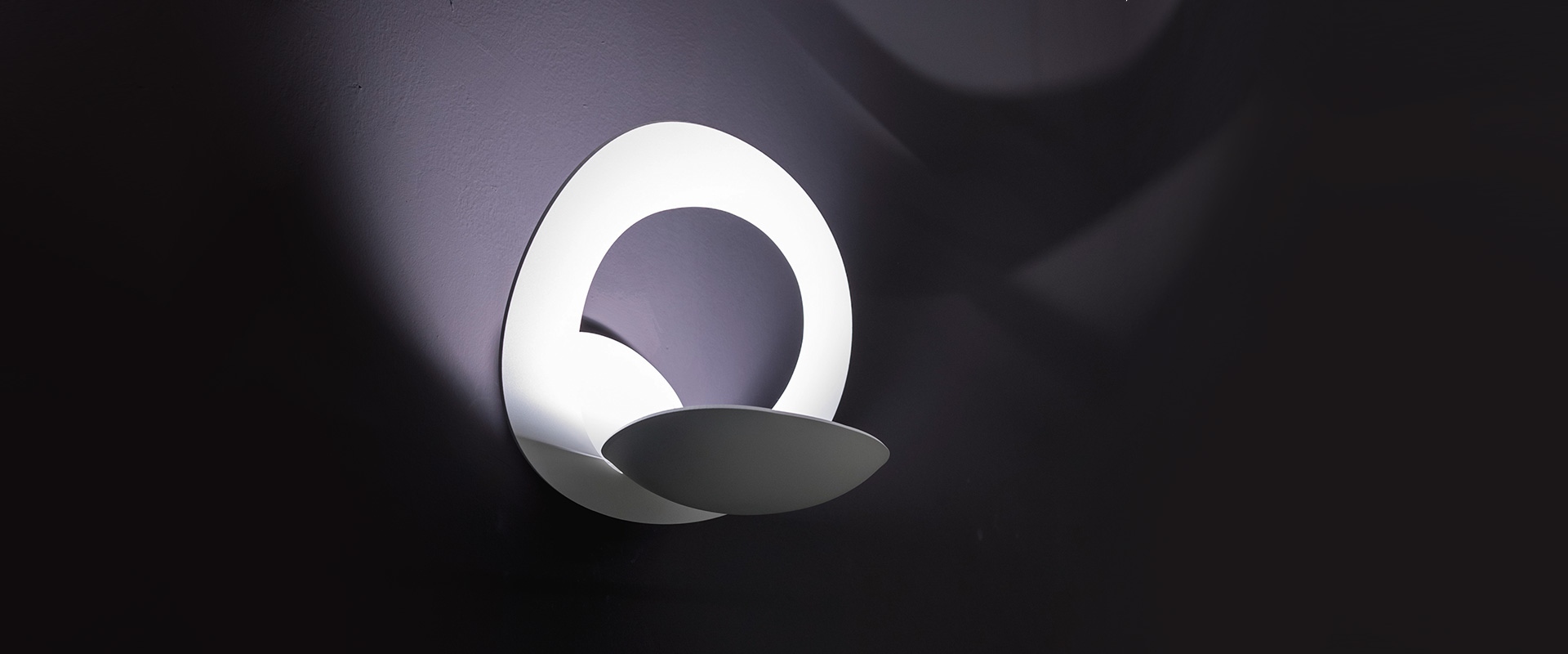 Artemide Pirce Parete wall lamp: sculptural white circular LED ring with curved lower reflector, casting dramatic shadows on dark wall