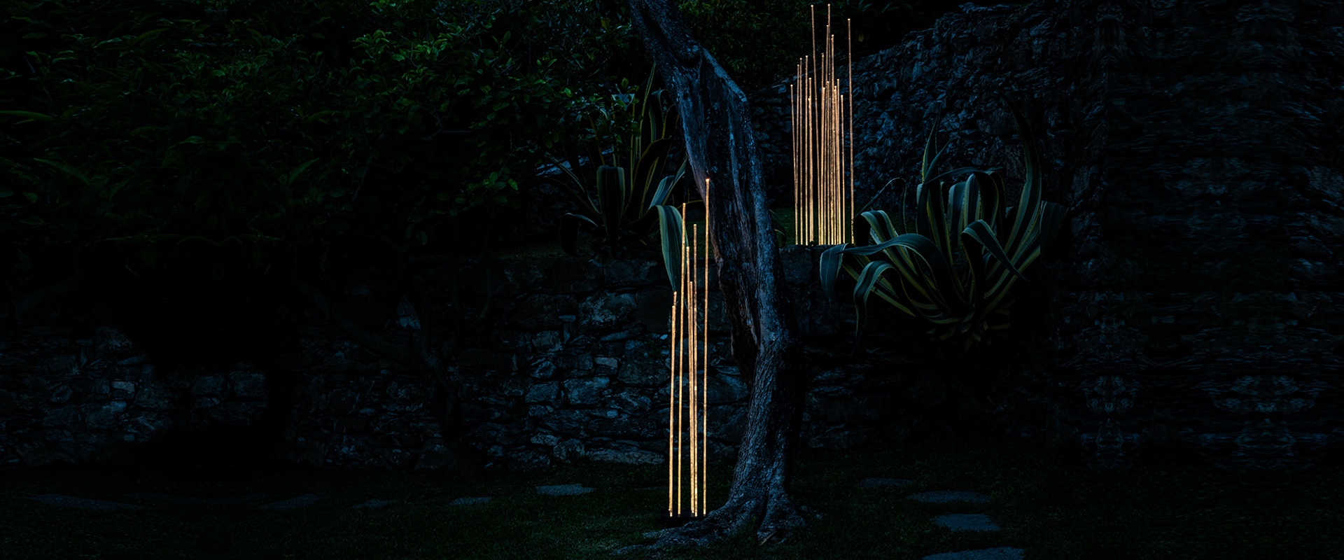 Artemide Reeds Outdoor floor lamp: cluster of illuminated vertical white rods glowing at night against red flowers backdrop