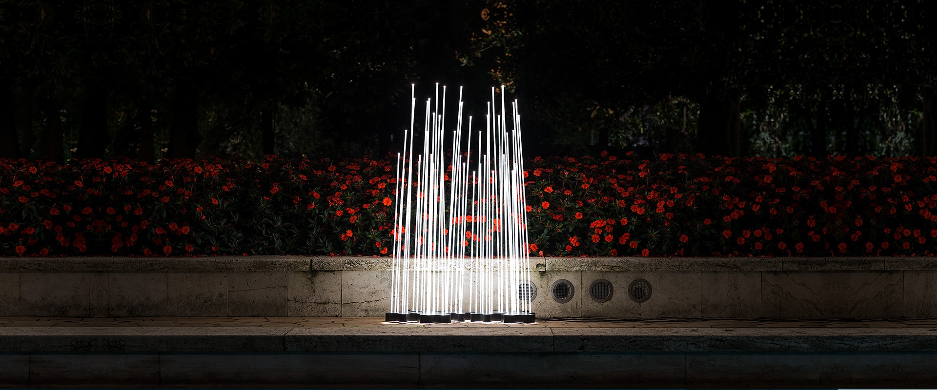Artemide Reeds Outdoor floor lamp: multiple illuminated vertical golden rods arranged like reeds, placed beside a tree in a dark garden