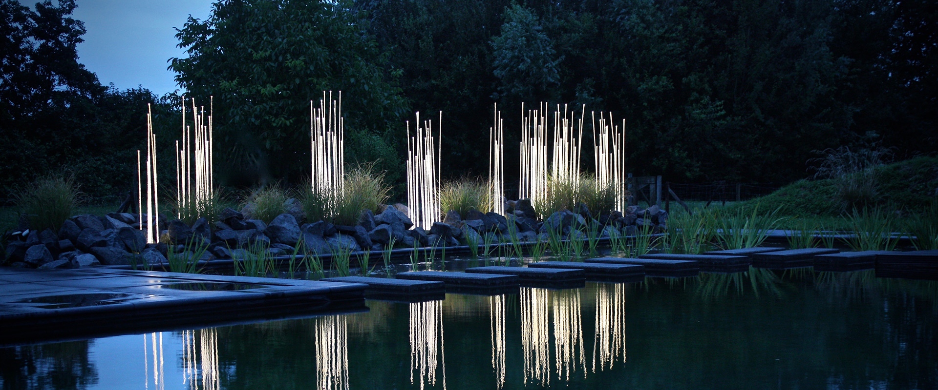 Artemide Reeds Outdoor luminaires: tall slender light columns reflected in a still pond, surrounded by lush garden vegetation at dusk