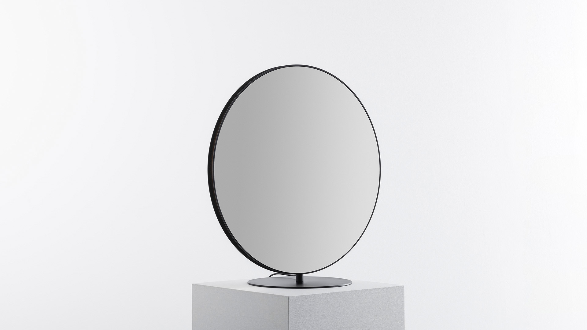 Round mirror on a stand reflecting a hand reaching toward it, with another hand visible in the background against a white wall.