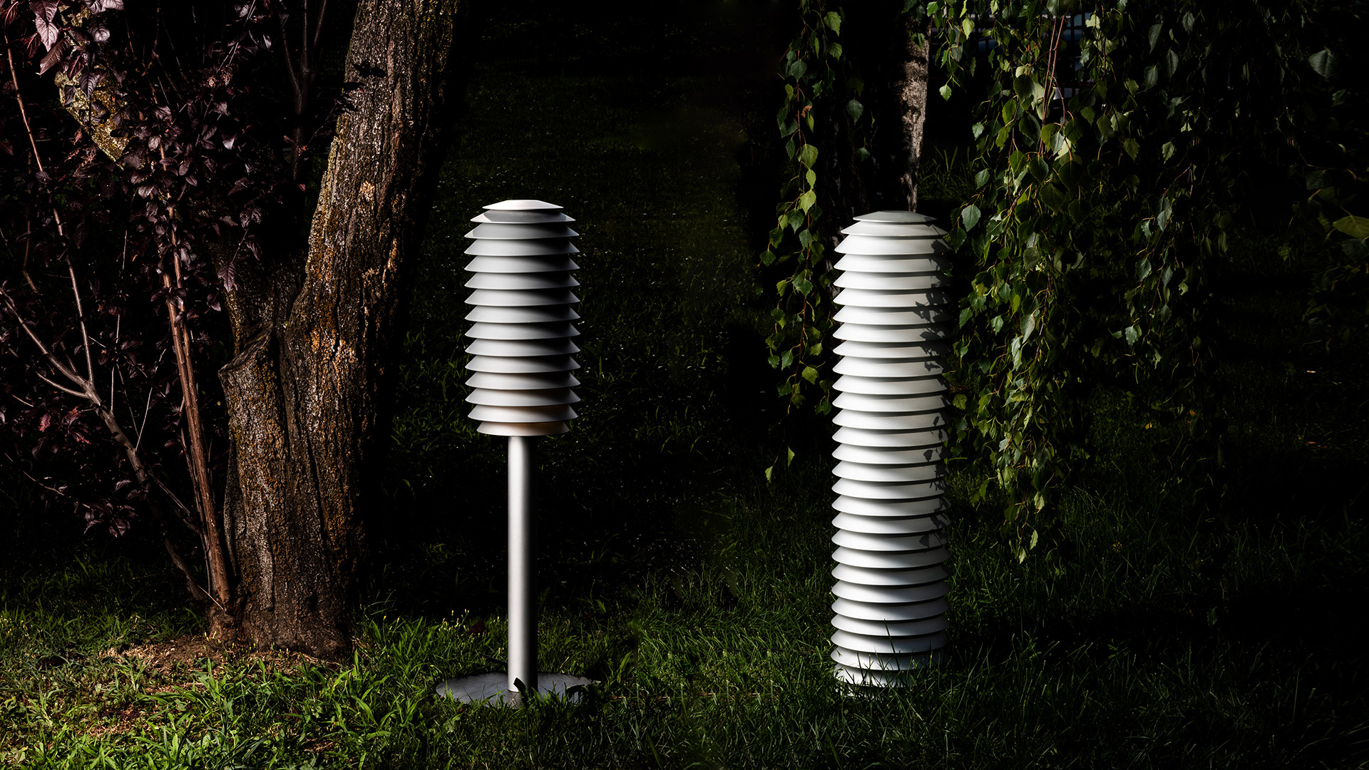 Two Artemide Slicing outdoor lamps with white stacked disc shades: one on a pole, one floor-standing, illuminating a garden at night