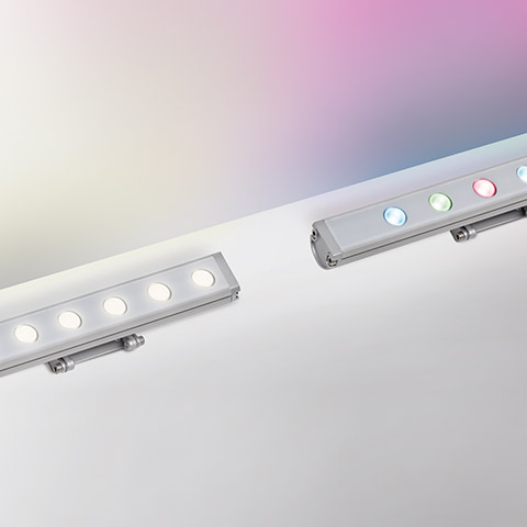 Two Artemide Spike LED bar fixtures: left emitting warm white light, right displaying multicolor RGB LEDs, both in aluminum housing