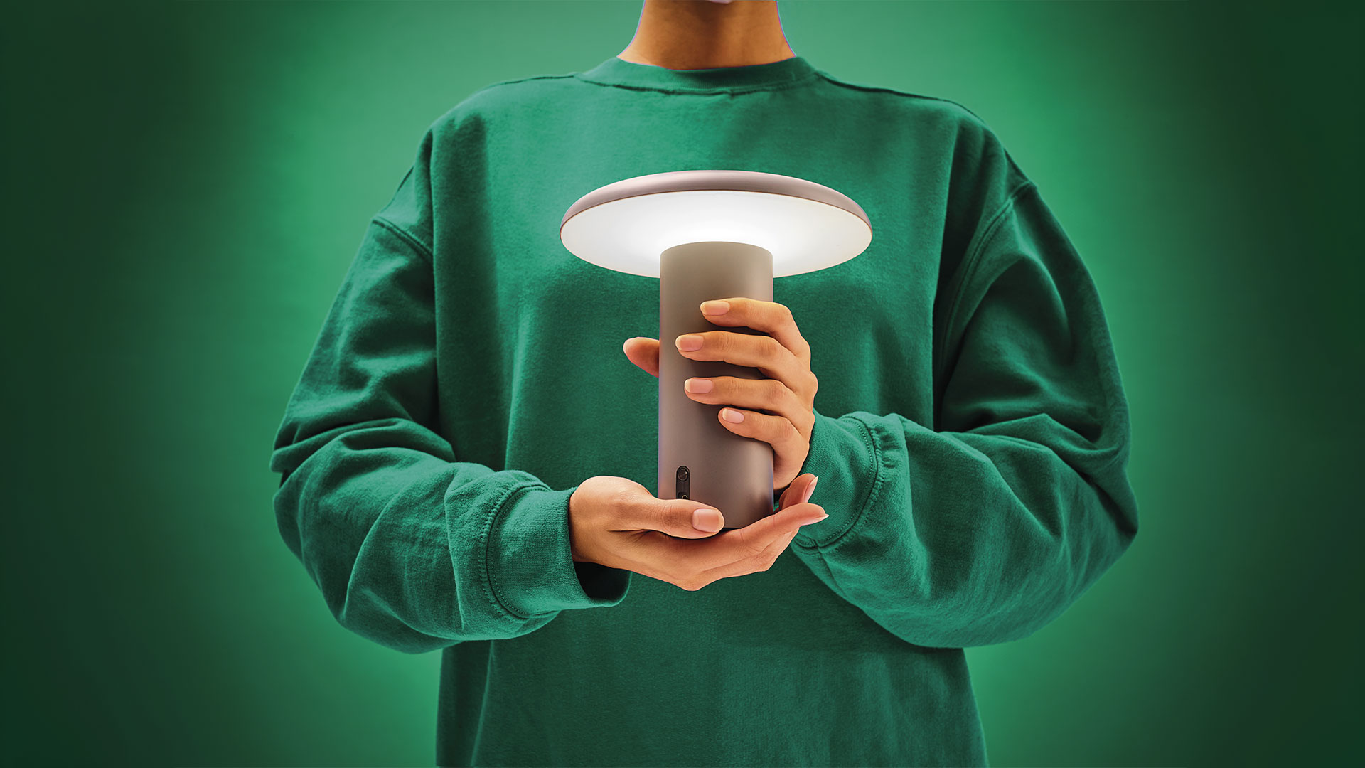 Hand holding Artemide Takku portable green lamp with cylindrical base and wide flat diffuser emitting warm light
