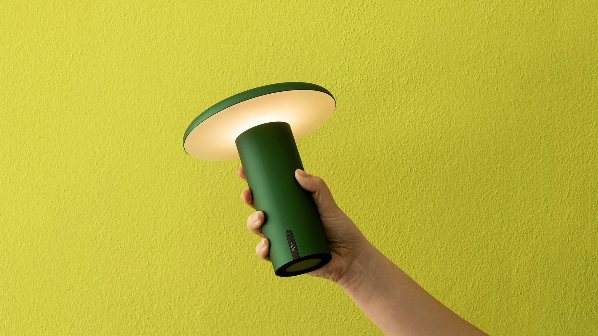 Hand holding Artemide Takku portable LED lamp with cylindrical grey body and flat circular diffuser emitting warm white light