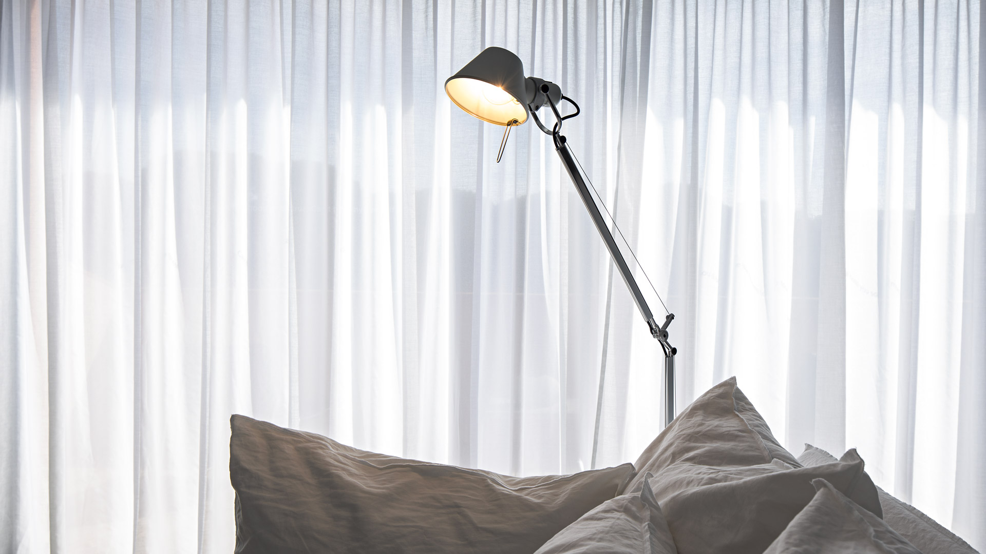 Artemide Tolomeo Terra floor lamp with dark shade and articulated arm, positioned beside pillows against sheer white curtains