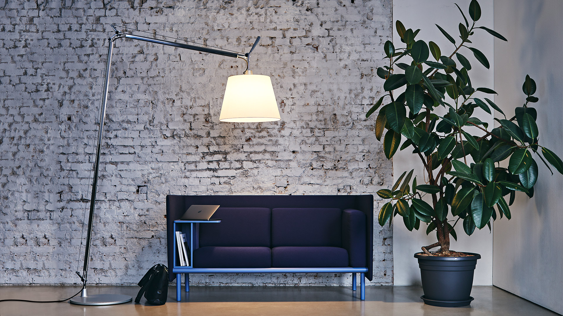 Artemide Tolomeo Maxi floor lamp with white shade illuminating a dark sofa against a white brick wall with a potted plant