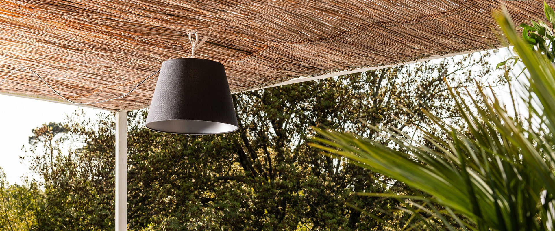 Tolomeo Paralume Outdoor pendant lamp with black conical shade hanging from a natural reed pergola, surrounded by lush greenery