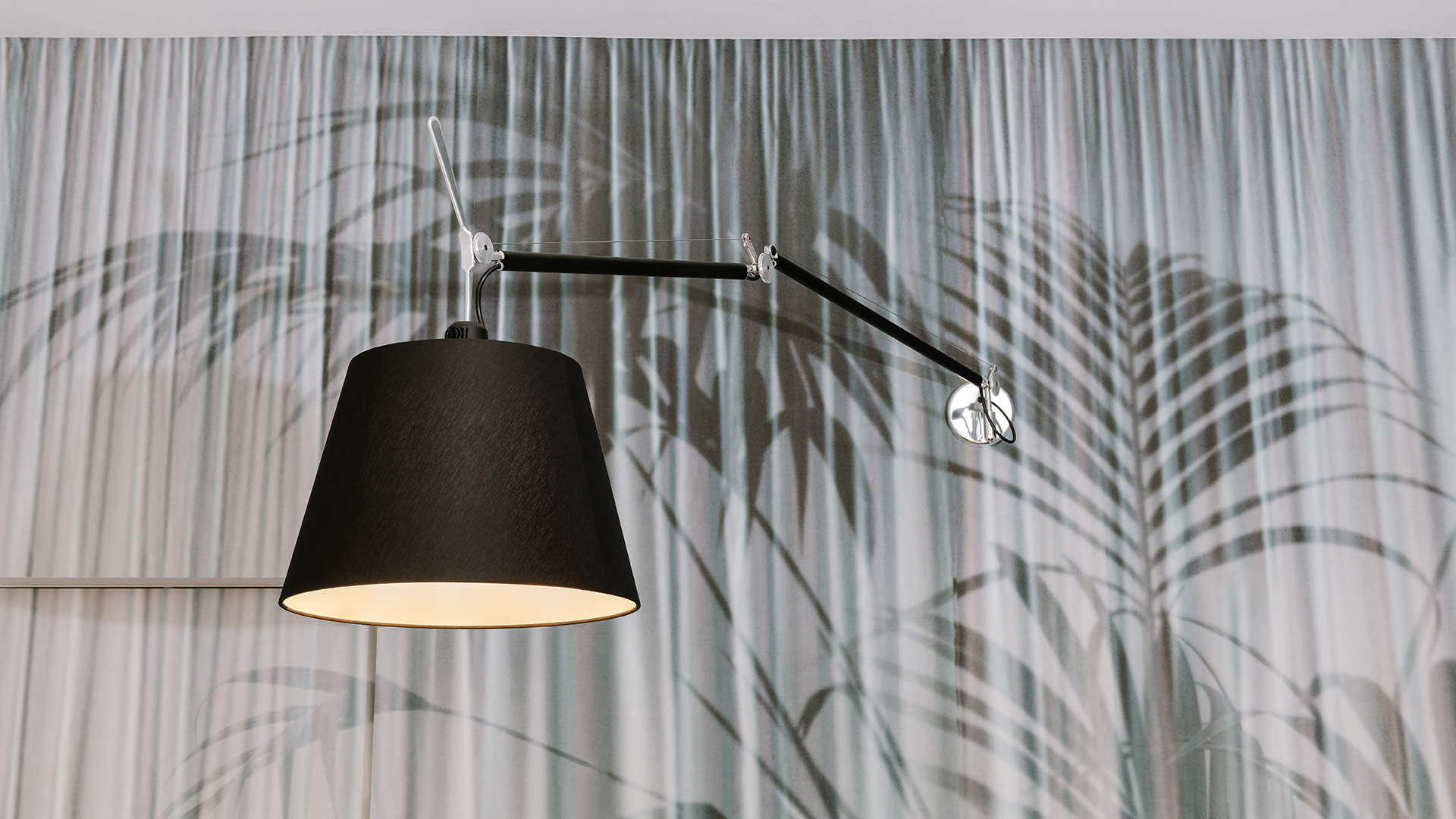 Artemide Tolomeo Mega Parete wall lamp with black fabric shade and adjustable aluminum arm, mounted against a tropical leaf-patterned curtain