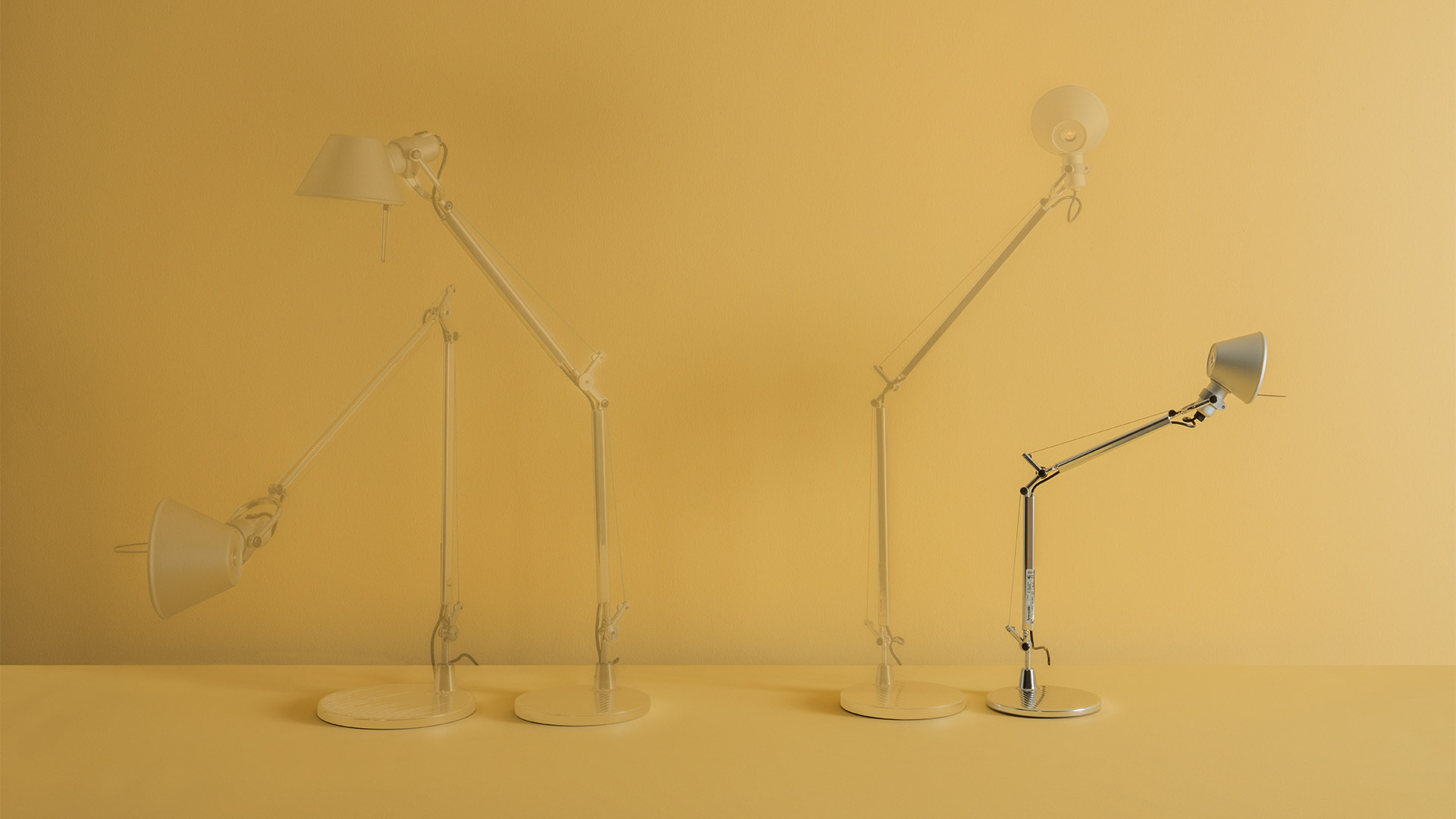 Four Artemide Tolomeo desk lamps in vibrant colors: blue-green, cobalt blue, yellow, and lavender, displayed on a white surface