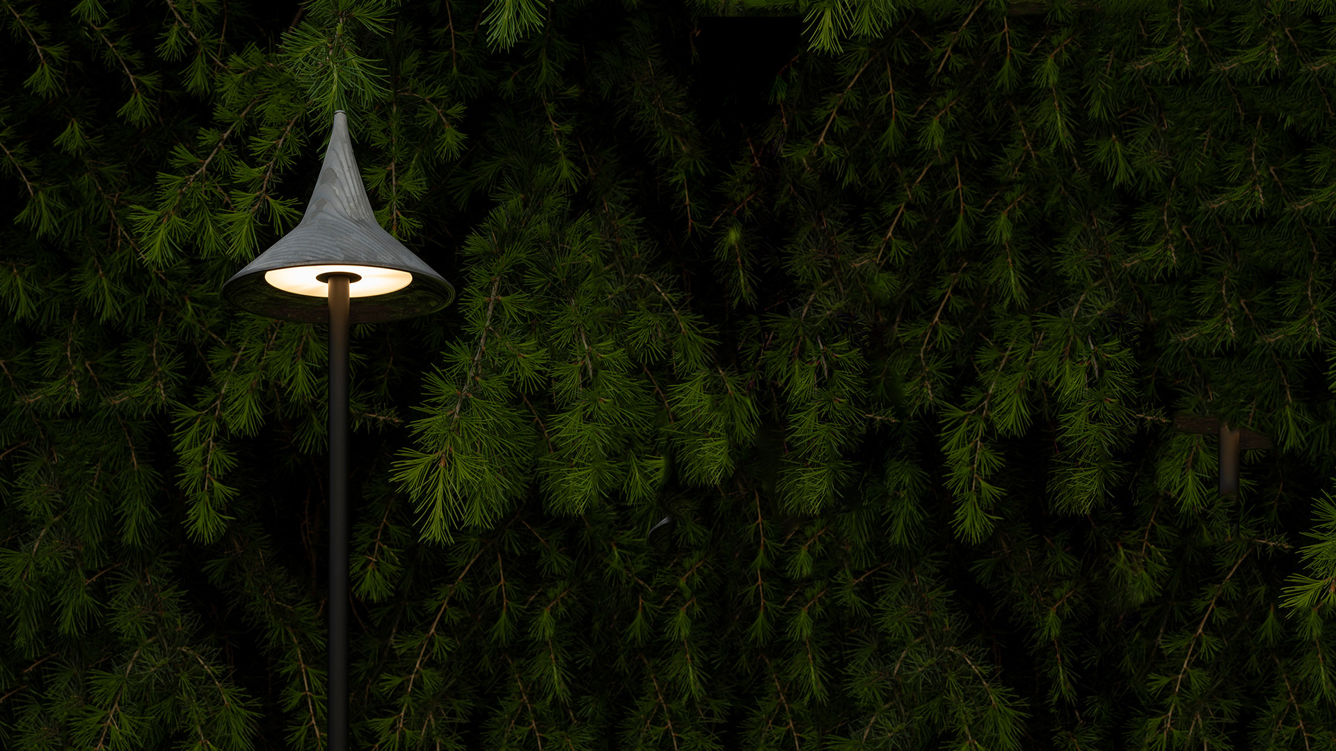 Artemide Unterlinden Outdoor pole lamp with conical shade emitting warm light, set against a dense evergreen hedge at dusk
