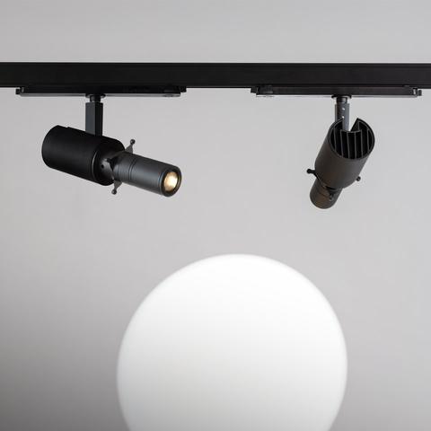 Two Artemide Vector black track spotlights mounted on a rail, one with a shaper accessory, projecting a circular beam of light